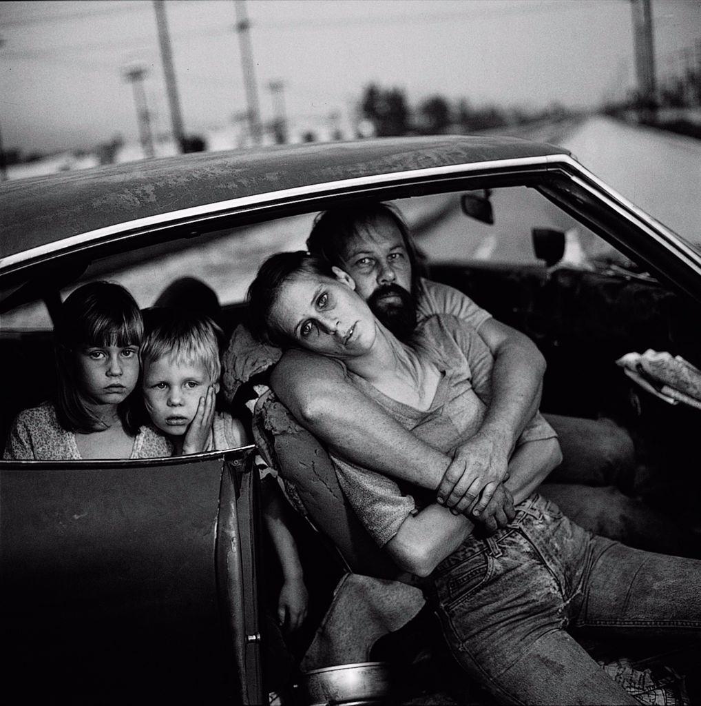 A stark black-and-white photograph of a homeless family inside their car. The mother leans into the father’s arms, her hollow-eyed expression conveying exhaustion. Two children sit in the car’s backseat, staring directly at the camera, their faces filled with uncertainty. The worn-down interior of the vehicle highlights the family's dire circumstances and the harsh realities of poverty.