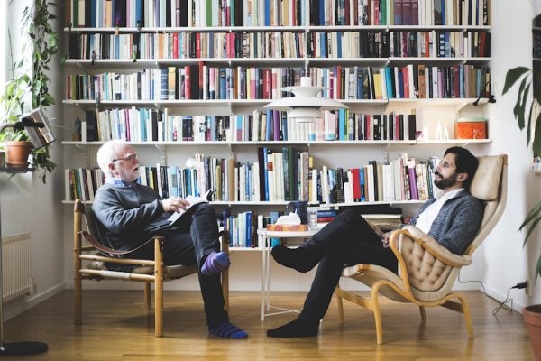 Therapist communicating with man while sitting by book shelf at home office