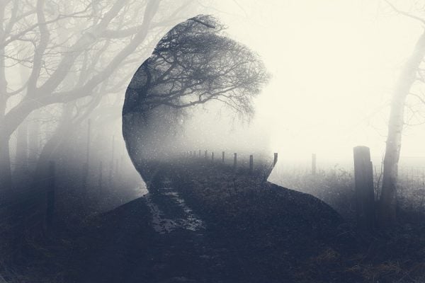 A double exposure of a spooky half transparent hooded figure layered over a foggy path in the countryside