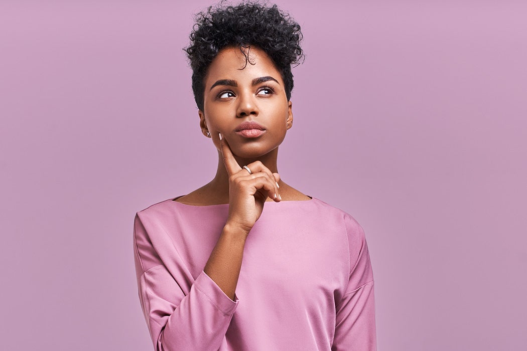 Pensive attractive curly African American female being deep in thoughts, raises eye, wears fashionable clothes, stands against lavender wall.