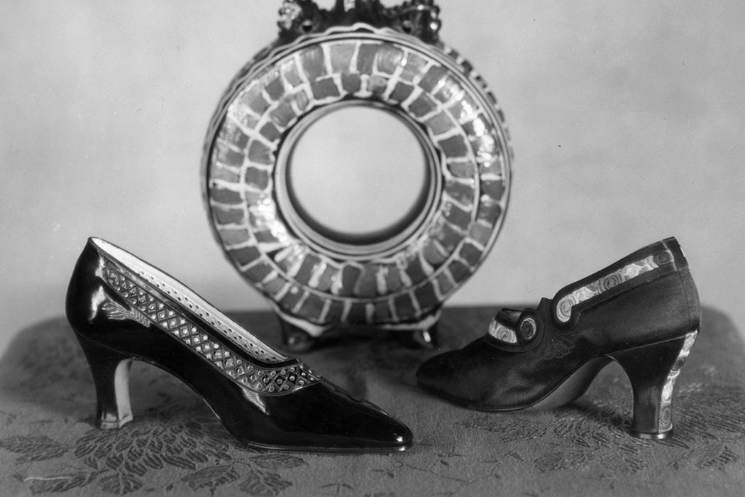 On the right high heeled black leather shoes with a colourful hand sewn design. On the left a patent leather high heeled court shoe with a perforated trim, c. 1925