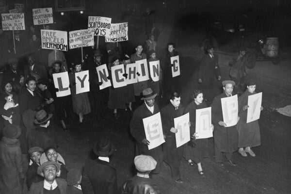 A high-angle view of a protest march with protesters carrying banners and placards reading 'Lynching is a Social Blot, Wipe it Out!', 'Free the Scottsboro Boys', 'Free Angelo Herndon', and 'Lynching is Un-American, Stop! Lynching' with some of the protestors carrying individual letters that spell out 'Stop! Lynching', United States, circa 1934.