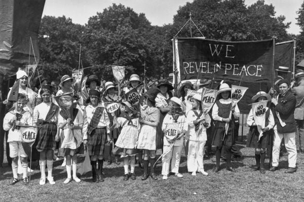 A group of children, among them are some dressed in Highland regalia with others wearing sailor suits, during League of Nations Rally in Hyde Park London, England, June 1921. The children, all of Hampstead, hold small signs reading 'Peace' and 'No War' and are gathered before a large banner reading 'We Revel in Peace'.