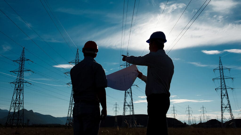 Two engineers in hardhats looking at high-voltage electrical lines on towers.